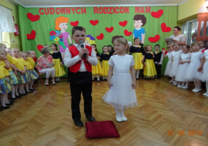 Dziewczynka w białej sukience oraz chłopiec w białej koszuli, czarno-czerwonej kamizelce oraz czarnych spodniach witają zebranych gości.